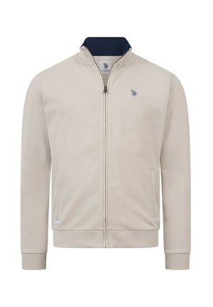 Beige Reißverschlussjacke mit gerippten Bündchen und Kragen, marineblauem Innenkragen, zwei Seitentaschen und kleinem, blau besticktem Logo auf der linken Brust.