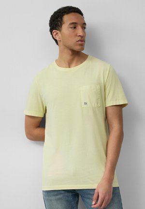 Man in een lichtgele T-shirt met korte mouwen en een borstzak, en blauwe jeans, staande tegen een effen lichtgrijze muur, die naar rechts kijkt.