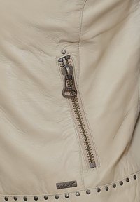 Der Abschnitt der beigen Lederjacke verfügt über einen silbernen Reißverschluss, dekorative Nieten am Saum und ein weiches, strukturiertes Material. Ein Logo-Tag ist vorhanden.