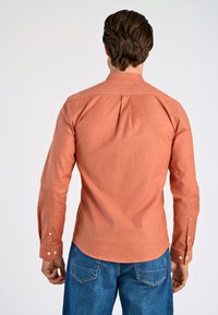 Camisa de color óxido hecha de tejido texturizado, con mangas largas y puños abotonados, y un pliegue en la parte de atrás. Combinando con jeans de mezclilla azul.