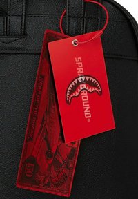 Bolsa negra texturizada con una etiqueta roja que presenta un gráfico de un tiburón y el texto "AÑO DEL TIBURÓN". La etiqueta está unida con un hilo rojo.