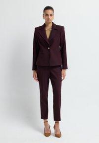 Blazer bordeaux avec un seul bouton doré et deux poches avant, associé à un pantalon bordeaux slim et des escarpins à bride cheville marron.