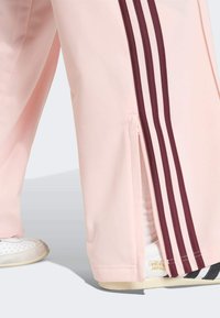 Różowe spodnie sportowe z bordowymi paskami, wyposażone w suwaki przy mankietach. Tkanina wydaje się gładka, o przewiewnej strukturze. Widoczne białe trampki.
