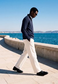 Homme portant des lunettes de soleil, un pull bleu marine, un pantalon blanc et des chaussures noires, marchant le long d'une promenade en bord de mer courbée par une journée ensoleillée.