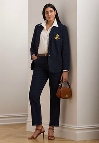 Marineblå blazer med gullknapper og emblem, hvit skjorte, mørke jeans, brunt belte, brun håndveske og brune sandaler med hæl. Glatte teksturer, skreddersydd passform.