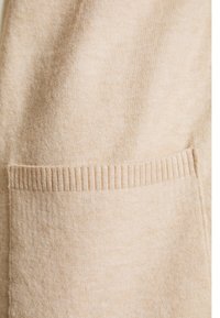 Tessuto beige con una superficie liscia e un dettaglio di tasca a righe. Il design complessivo è semplice e privo di ornamenti.