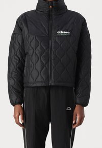 Czarna pikowana kurtka z wysokim kołnierzem, z zamkiem błyskawicznym z przodu i logo. Teksturowany wzór i krótki fason, połączona z czarnymi spodniami.