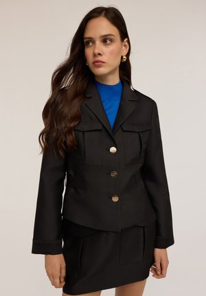 Blazer negro con botones dorados, dos bolsillos frontales y una forma estructurada, combinado con un cuello de tortuga azul. Tejido suave y mate.