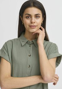Camicia button-up verde salvia con colletto, maniche corte arrotolate e bottoni decorativi. Tessuto dalla trama liscia, indossata da un modello con la mano sul mento.