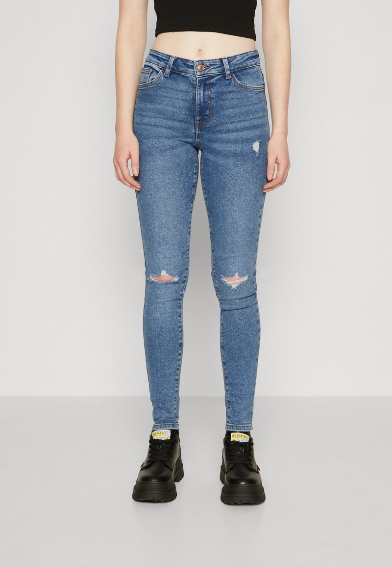 Jeans skinny bleus avec détails déchirés aux genoux, fabriqués en denim, design taille haute, associés à des chaussures noires à semelles épaisses.