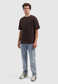Donkerbruine t-shirt met korte mouwen en een borstzak, gecombineerd met lichtblauwe distressed jeans en metallic sneakers. Neutrale achtergrond.