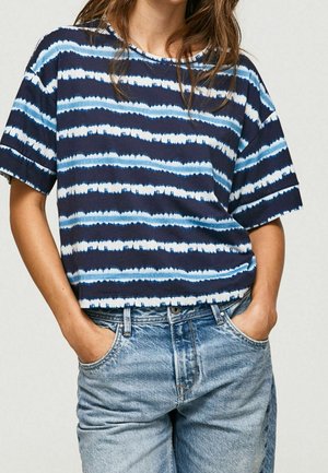 Žena oblečená v modrobílém pruhovaném tričku s krátkým rukávem, zasunutém do světlých džínů, ruce v kapsách.