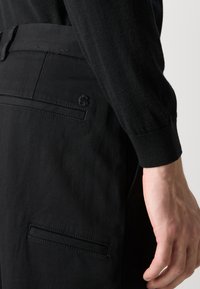Black trousers with a smooth texture, featuring a rear pocket and subtle branding. The waistband is flat, and the fabric appears lightweight.