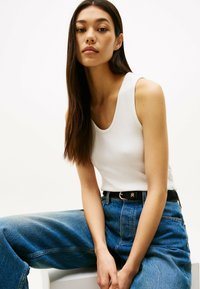 Weißes geripptes Tanktop kombiniert mit hochtaillierten, weiten blauen Jeans. Schwarzer Gürtel mit einer goldenen Schnalle. Glatte Texturen mit Farbkontrasten.