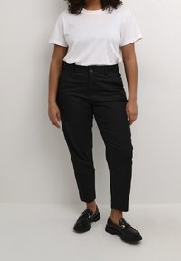 Pantalon noir sur mesure à texture lisse, jambe fuselée, poches avant et fermeture par bouton. Associé à un t-shirt en coton blanc et des mocassins noirs.