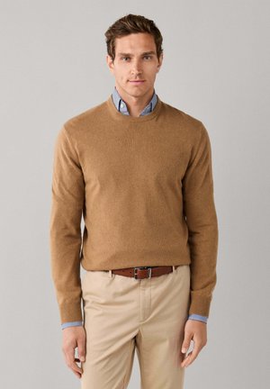 Man with short brown hair wears light brown sweater over blue striped shirt and beige pants with brown belt, standing against plain gray background.