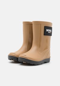 Bottes en caoutchouc imperméables de couleur beige avec un patch logo noir, semelle à grip texturé et finition lisse, présentant une forme arrondie et une hauteur courte.