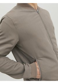 Homme portant une veste zippée marron clair avec la main dans la poche latérale zippée sur fond uni.