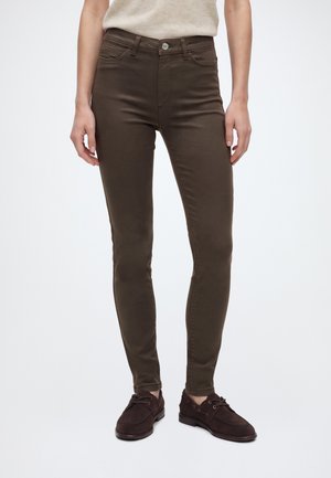 Person wearing dark brown slim-fit pants and dark brown lace-up shoes, standing against a plain light background.
