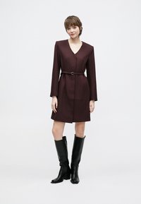 Μακρυμάνικο μαρεν φόρεμα με λαιμόκοψη V, ζώνη στη μέση. Συνδυασμένο με μαύρες δερμάτινες μπότες μέχρι το γόνατο. Απλός, δομημένος σχεδιασμός.