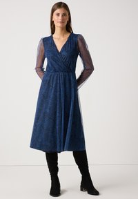 Blaues Wickelkleid mit floralem Muster, langen transparenten Ärmeln und V-Ausschnitt. Gestylt mit kniehohen schwarzen Stiefeln und einer fließenden Silhouette.