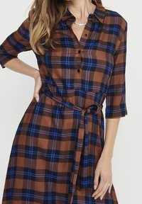 Abito camicia a quadri marrone con accenti blu e neri, chiusura frontale con bottoni, maniche a tre quarti e dettaglio in vita legato. Tessuto dalla consistenza liscia.