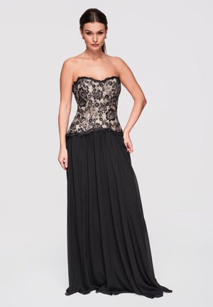 Mujer con corsé negro de encaje sin tirantes y falda larga negra y fluida, de pie con la mano en la cadera contra un fondo liso.