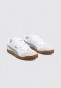 Zapatillas blancas en materiales de ante y cuero liso, con una franja lateral gris, punta redonda y suela de goma marrón.