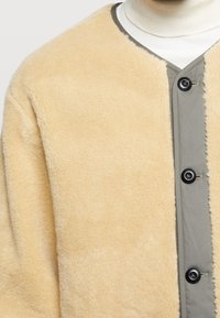 Giacca in shearling beige con una chiusura frontale grigia, caratterizzata da un tessuto testurizzato e una finitura morbida, progettata con un colletto rotondo.
