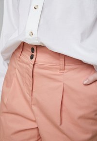 Pantalones cortos de algodón rosa claro con corte recto, con dos botones negros y pliegues sutiles en la parte delantera, combinados con una camisa blanca con cuello.