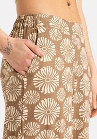 Pantaloni maronii cu un model floral alb de mari dimensiuni, cu un talie elastică și buzunare laterale. Textura pare ușoară și respirabilă.