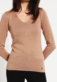Beige långärmad V-ringad tröja gjord av ribbade tyger. Har en subtil logodetalj nära fållen, figurnära silhuett och mjuk textur.
