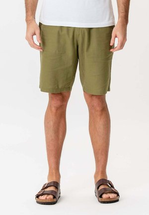Mand iført olivengrønne shorts, en hvid skjorte og brune remsandaler, der står foran en ensfarvet baggrund.