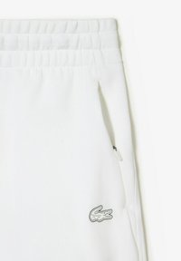 Pantaloni della tuta bianchi realizzati in tessuto morbido, con elastico in vita, tasca con zip e un piccolo logo del coccodrillo Lacoste in outline nero.