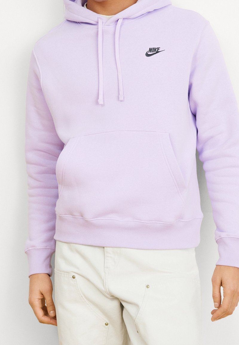 Person som bär en ljuslila Nike-hoodie med en framficka och beige byxor, står mot en vit bakgrund.