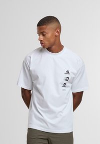 Joven con cabello corto, vistiendo una camiseta blanca oversized con logos negros en el pecho izquierdo, de pie contra un fondo claro y liso.