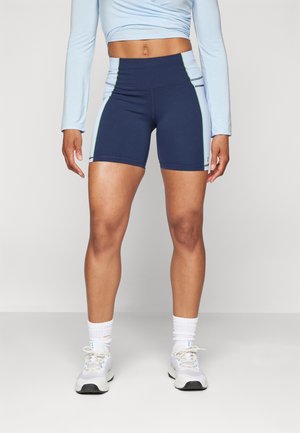 Vrouw draagt marineblauwe en lichtblauwe sportshorts, lichtblauwe crop top met lange mouwen, witte sokken en witte sneakers, staand tegen een effen achtergrond.