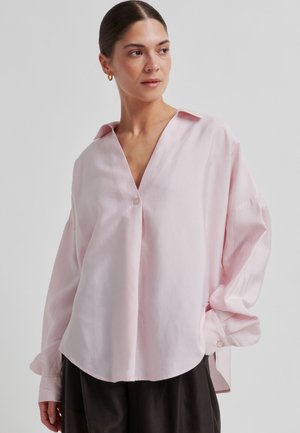 Jasnoróżowa, oversize koszula z dekoltem w kształcie litery V, zapinana na guziki, z bufiastymi rękawami. Wykonana z gładkiej tkaniny, z zaokrąglonym dołem.