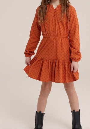 Enfant portant une robe orange texturée à manches longues avec des boutons et des bottines noires, debout les jambes écartées devant un fond uni.