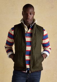 Junger Mann trägt eine dunkel gesteppte Weste über einem blau-roten, weiß gestreiften Langarm-Poloshirt mit hochgestelltem Kragen, steht vor beigem Hintergrund.