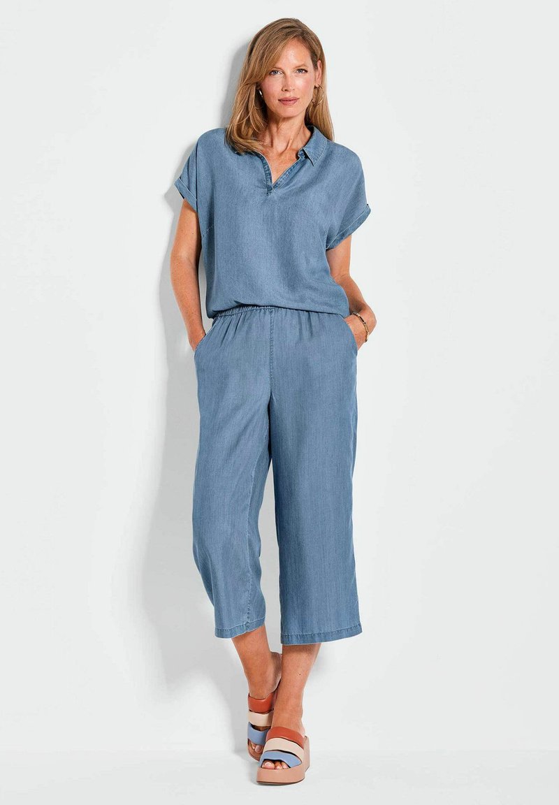 Denim-Overall in Hellblau, mit kurzen Ärmeln, Kragen, elastischem Bund und weit geschnittenen, kurzen Beinen. Getragen mit mehrfarbigen Plateausandalen.