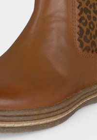 Bottine en cuir marron avec semelle texturée et superposée, et panneau latéral élastique imprimé léopard avec couture en zigzag sur les bords.