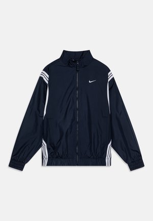 Jūrinis vėjo nepralaidus džemperis su užtrauktuku, aukšta apykakle, elastingomis rankogaliais ir baltomis šoninėmis juostelėmis. Ant krūtinės yra Nike logotipas.