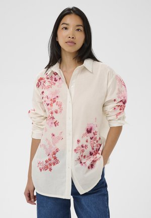 Young woman wearing a cream buttoned shirt with pink floral prints and blue jeans, standing with one hand in pocket against white background.