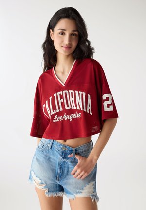 Młoda kobieta w czerwonym krótkim sportowym topie z napisem „California Los Angeles” i przetartych jasnoniebieskich dżinsowych szortach, stojąca na białym tle.