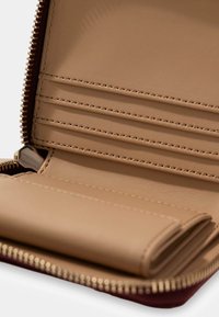 Portafoglio in pelle beige con chiusura a zip, scomparti interni per carte e finitura testurizzata. Presenta una base bordeaux a contrasto.
