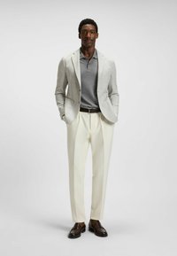 Jasnoszary marynarka w kratę noszona na szarym polo, w połączeniu z kremowymi spodniami i brązowymi loafersami. Posiada klasyczny kołnierz i akcent w postaci paska.