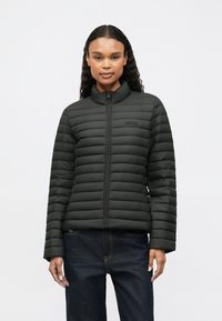 Femeie modelând o jachetă neagră matlasată cu fermoar și guler înalt, purtată cu blugi de culoare albastru închis, împotriva unui fundal alb simplu.