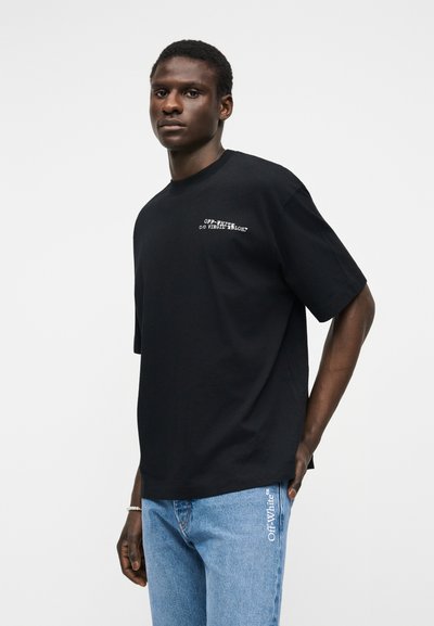 Jeune homme portant un t-shirt noir Off-White et un jean bleu avec le texte "Off-White" sur la jambe, posant devant un fond blanc uni.