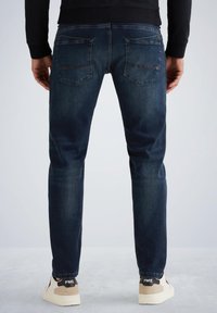 Jeans de mezclilla azul oscuro de corte slim, con cinco bolsillos y un ligero desgaste. La parte trasera cuenta con dos bolsillos de parche y un acento de logo.